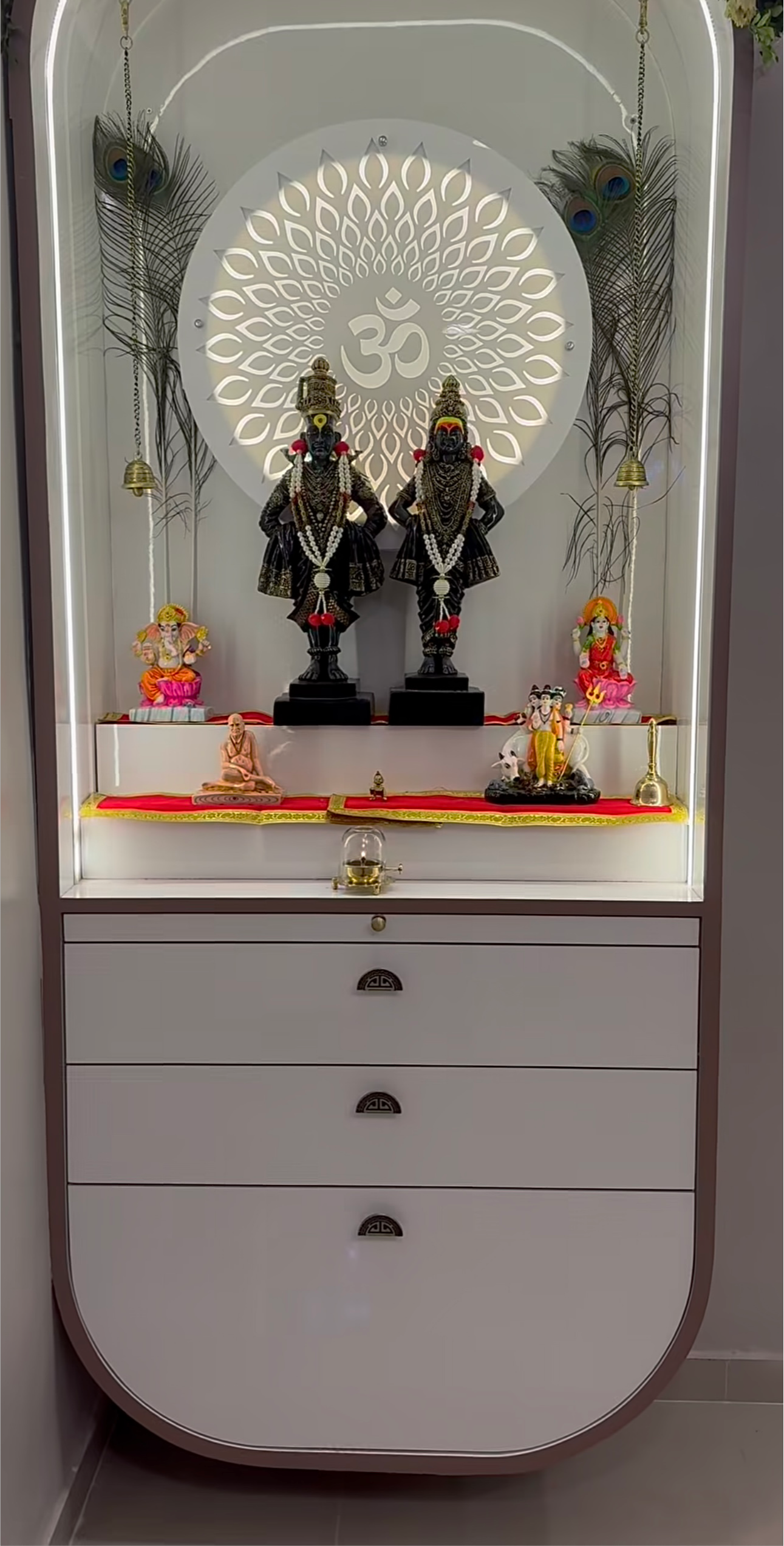 Contemporary white Pooja Mandir with rounded edges, backlit Om symbol, decorative flower arch, and traditional idols of Vithoba-Rukmini, Ganesh, and Lakshmi, perfect for modern Indian interiors.