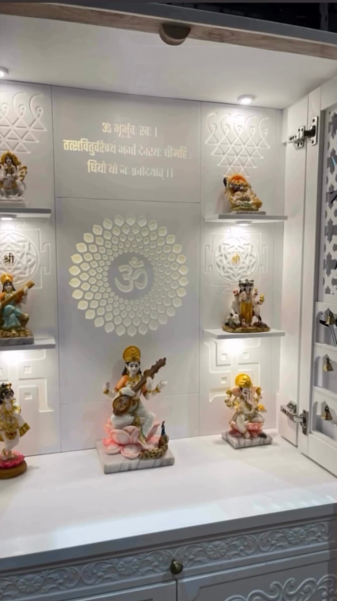 Modern white pooja mandir featuring decorative carvings, centered Om symbol, and elevated shelves displaying detailed idols of Hindu gods and goddesses with soft white lighting for divine ambiance.