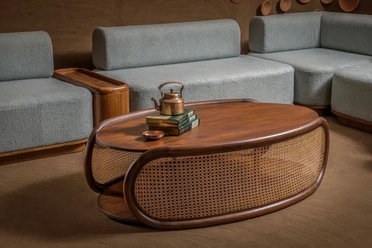 Mid-century modern oval wooden coffee table with rattan cane weaving, placed in a stylish living room with light gray sectional sofas and a warm-toned interior.