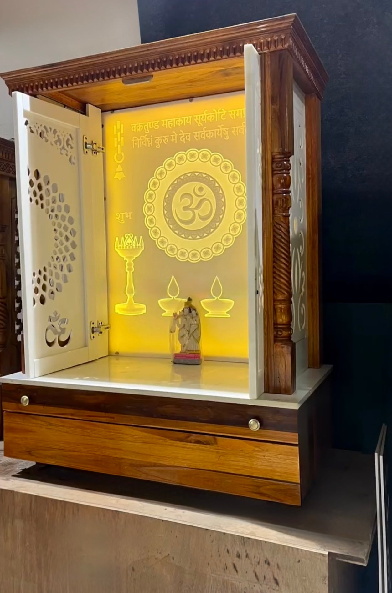 Handcrafted teakwood home temple with white Corian doors, backlit religious symbols, and elevated platform for deities.