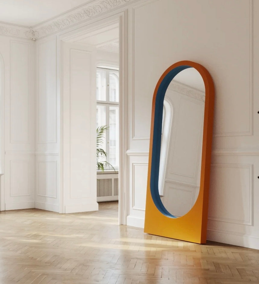 Contemporary orange and blue standing mirror placed in a bright, elegant interior with wooden flooring and decorative wall panels.