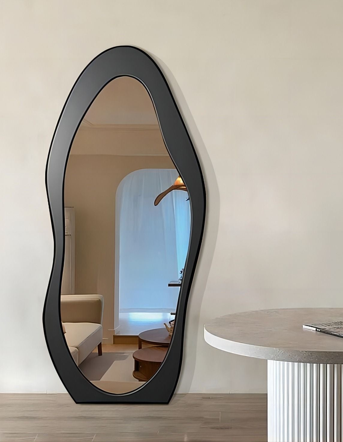 Irregular full-length mirror with black wavy frame leaning against a neutral-toned wall in a modern living room.