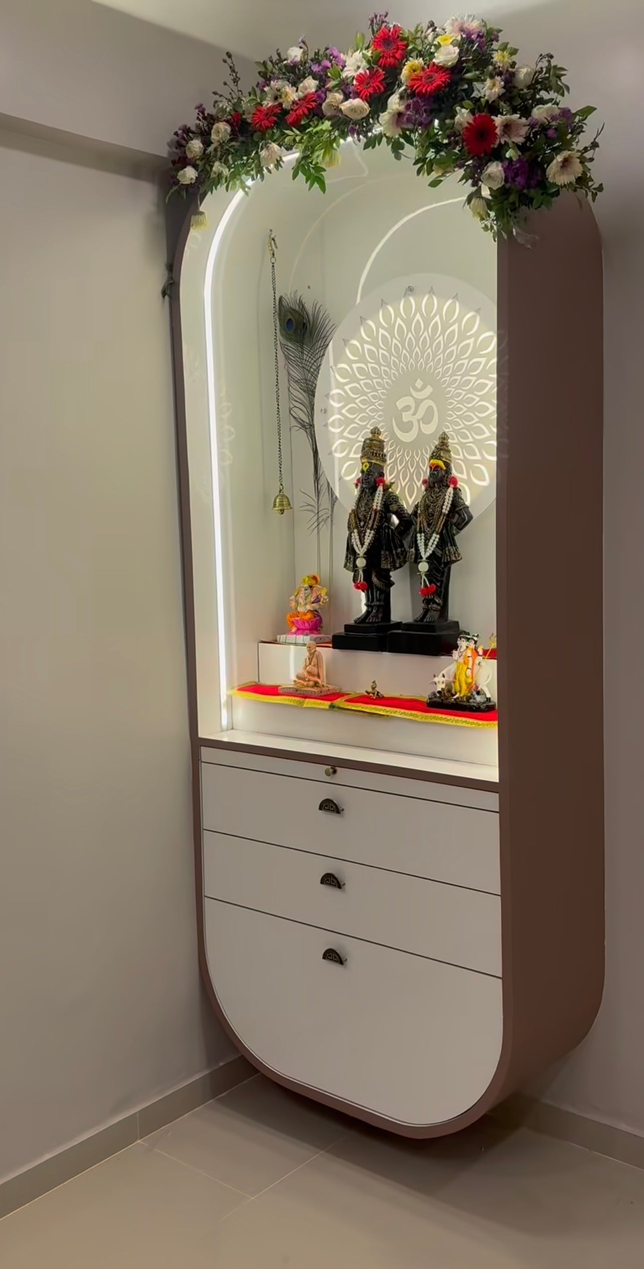 Modern curved-edge Pooja mandir for home with decorative lighting, peacock feathers, floral top arch, and traditional Hindu idols on a white altar.