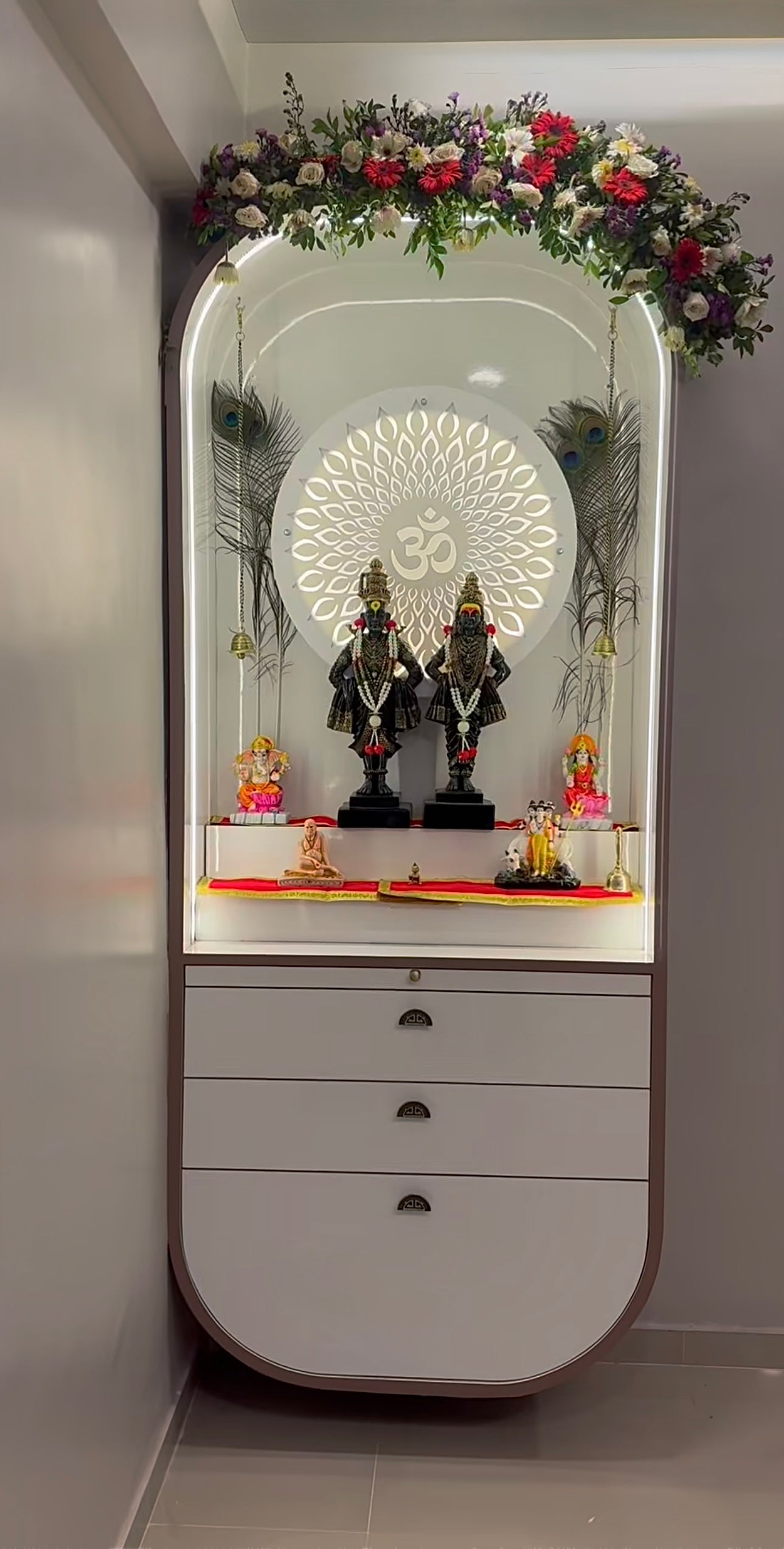 Premium wall-mounted mandir with backlit Om design, artificial flower garland arch, multiple deity idols, and pull-out drawer storage in a compact space.