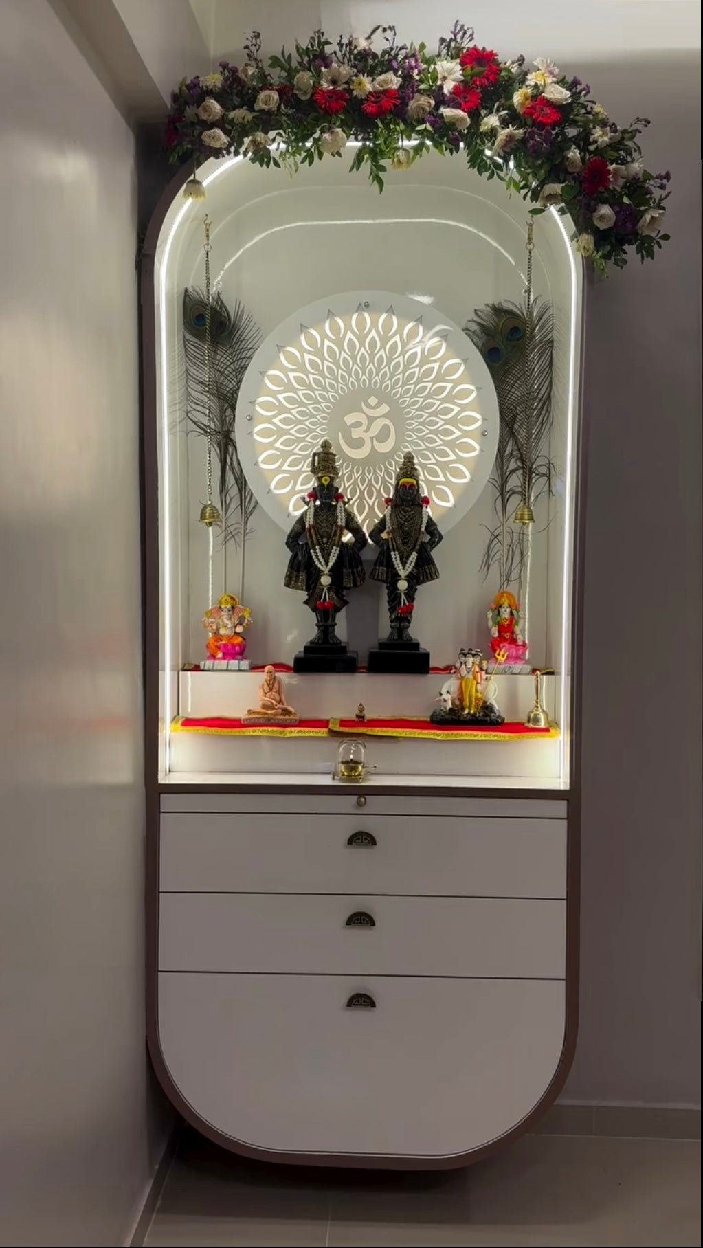 Elegant prayer altar for home with glowing Om backdrop, peacock feathers, brass bells, and neatly arranged Hindu deities.