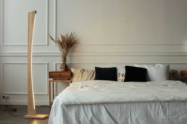 Modern curved wooden LED floor lamp with warm neutral light placed beside a bed and wooden nightstand in a minimalist bedroom.