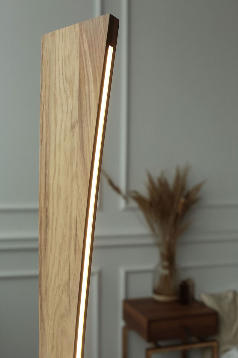 Close-up of a modern curved wooden LED floor lamp with warm white lighting, placed in a minimal living room interior with panelled wall and rustic decor.