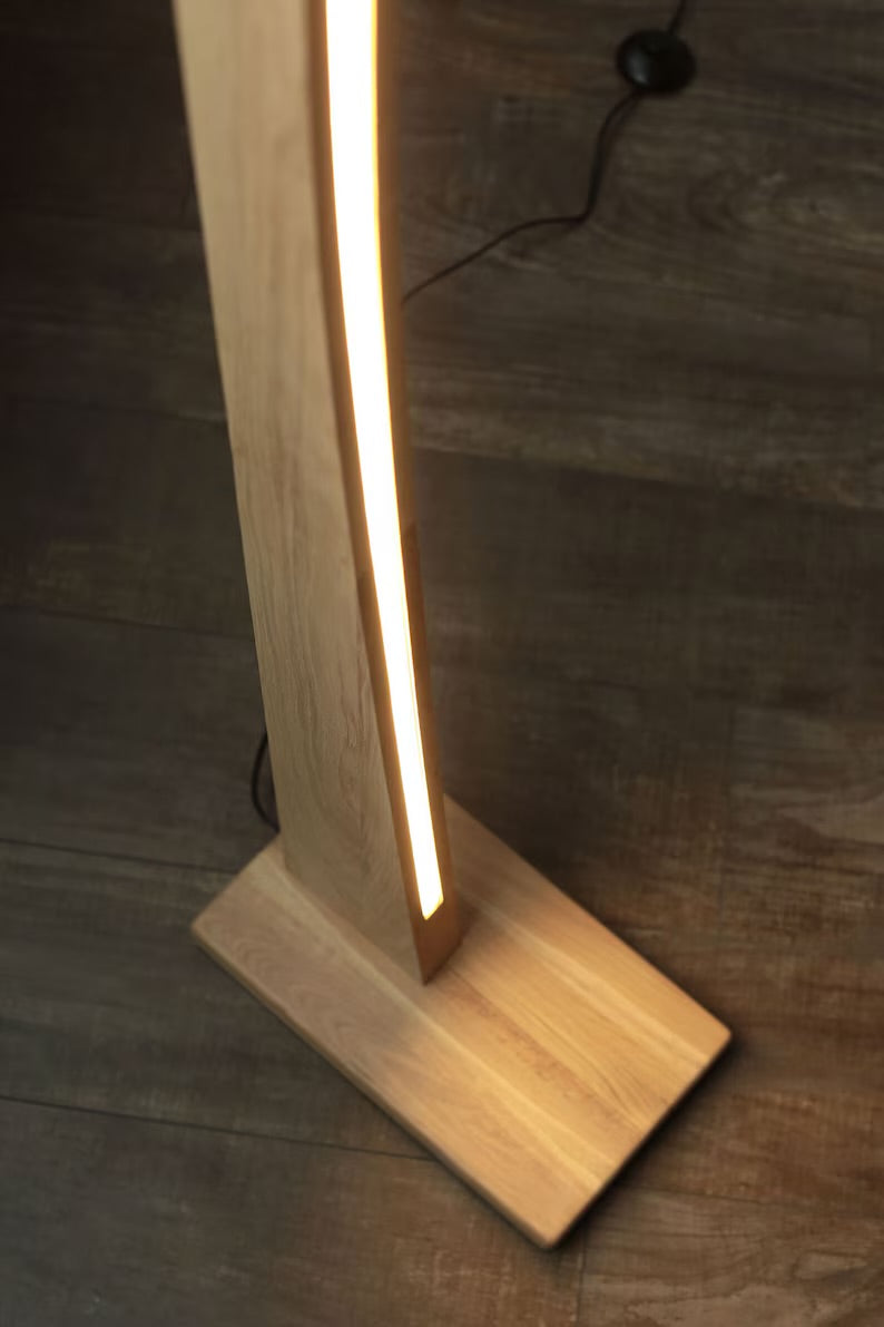 Close-up view of the wooden base and glowing LED strip of a modern curved floor lamp, placed on a dark wooden floor.