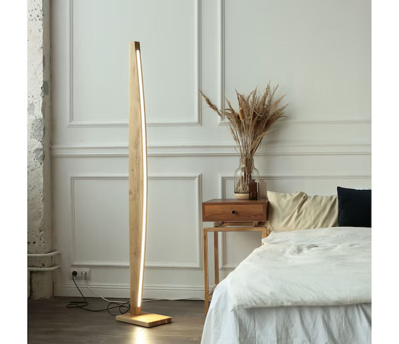 Sleek wooden LED floor lamp beside a bed and wooden side table with dried pampas in a vase, in a cozy modern bedroom.