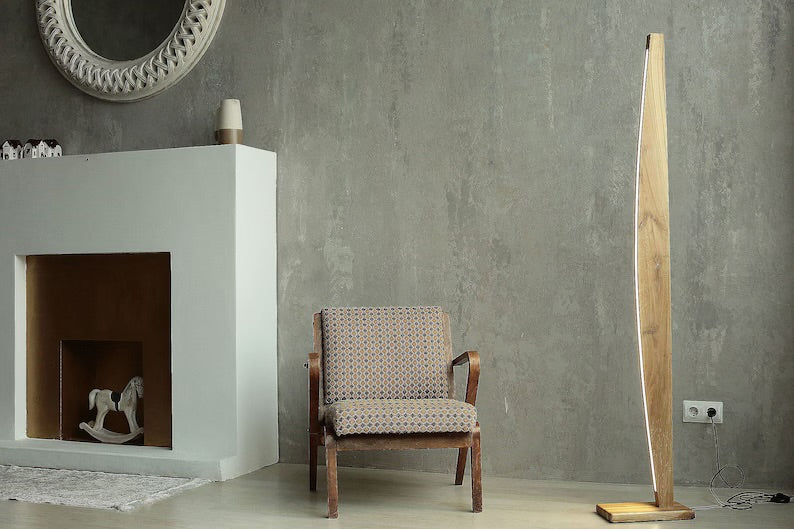 Modern wooden LED floor lamp with minimalist curved design, placed in a contemporary living room with grey textured walls, a patterned armchair, and a decorative white fireplace.