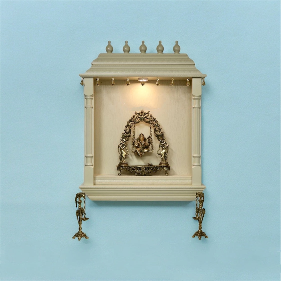 Wall-mounted pooja mandir against a pastel blue background, showcasing a brass Ganesha swing idol under warm spotlight with ornate side diya stands.