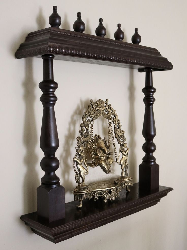 Wall-mounted dark wooden temple shelf with brass Ganesha idol on a swing, supported by intricately carved side pillars, ideal for home puja decor.