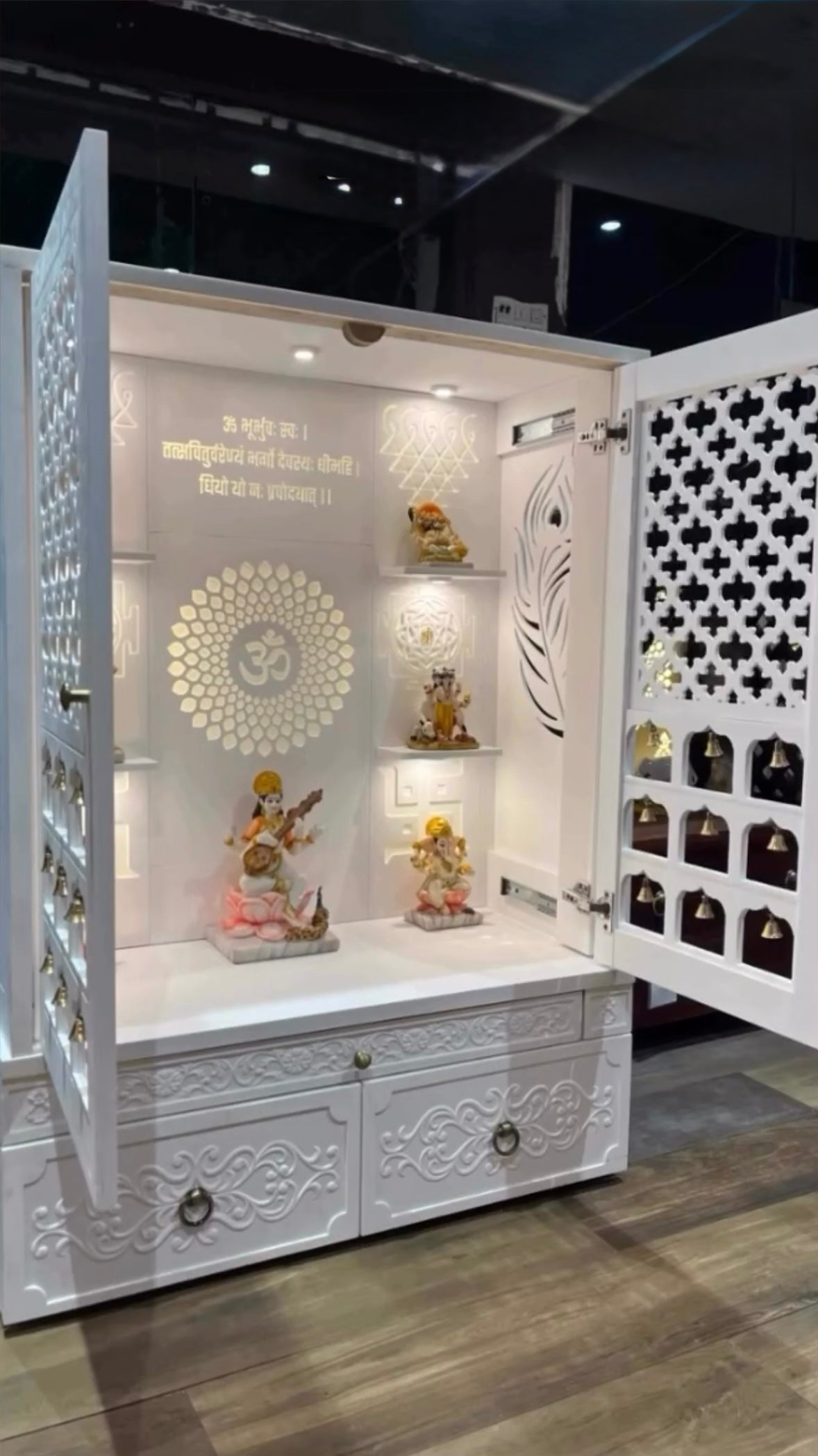 Contemporary handcrafted white temple for home with laser-cut jali door, brass bells, carved drawers, and a central sacred space adorned with religious idols and mantra wall art in golden finish.