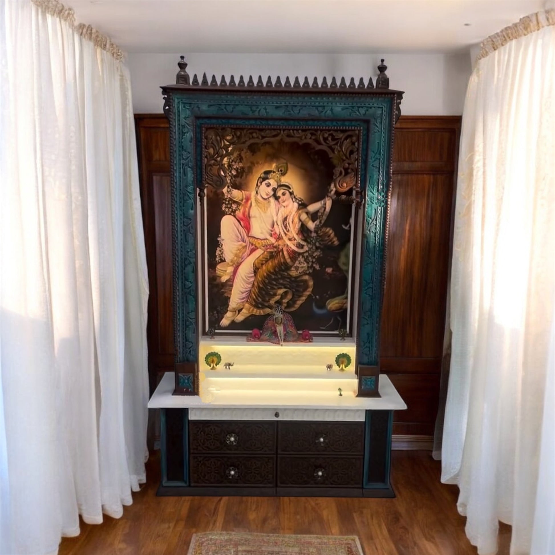 Elegant large wooden pooja temple in dark brown with vibrant peacock highlights, showcasing a Corian Radhe Krishna image and detailed drawer carvings.