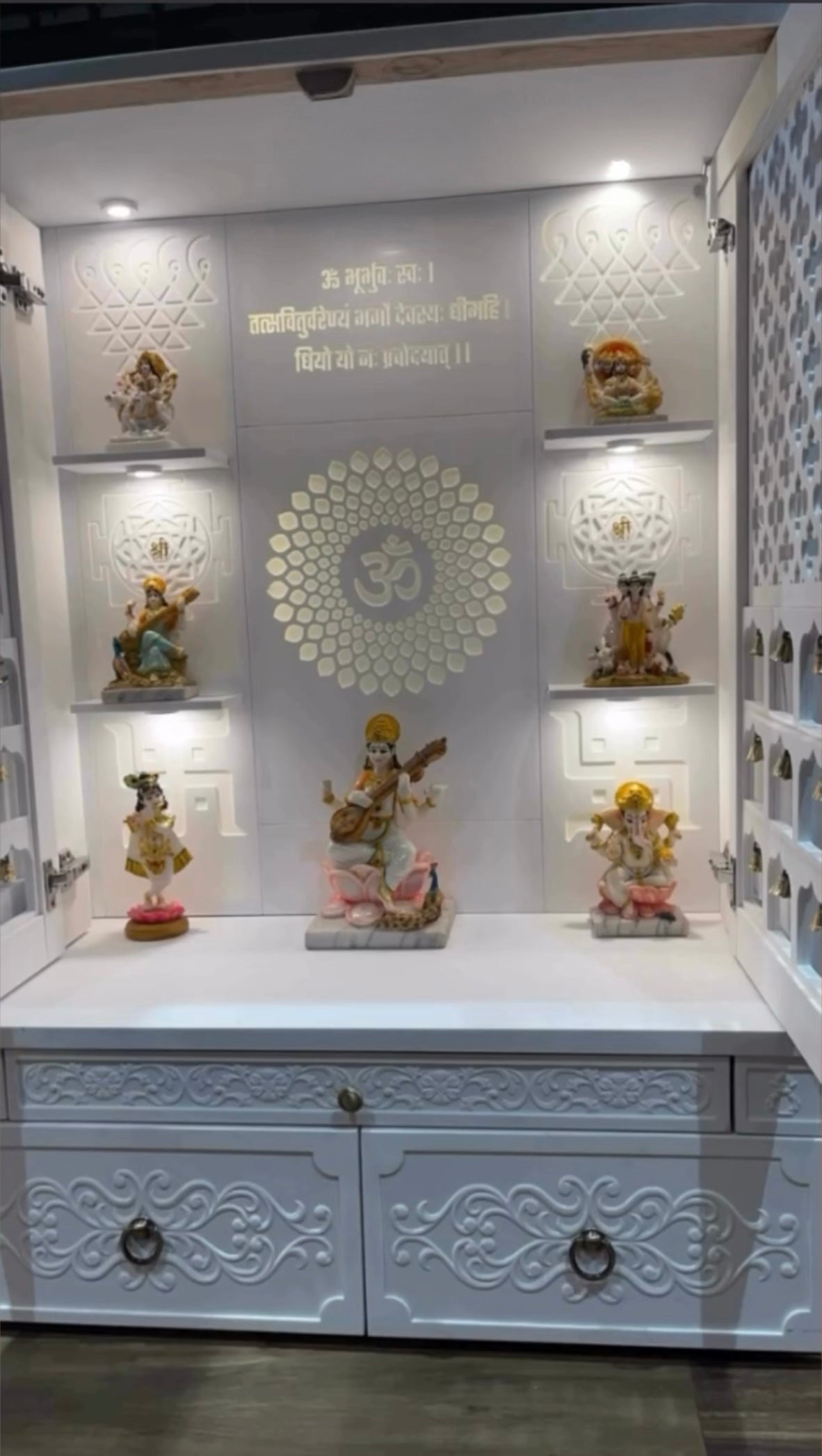 Elegant white wooden pooja mandir with intricate floral carvings, LED-lit shelves, and Hindu deity idols including Saraswati, Ganesh, Krishna, and Lakshmi, set against a sacred Om backdrop and Gayatri Mantra engraving.