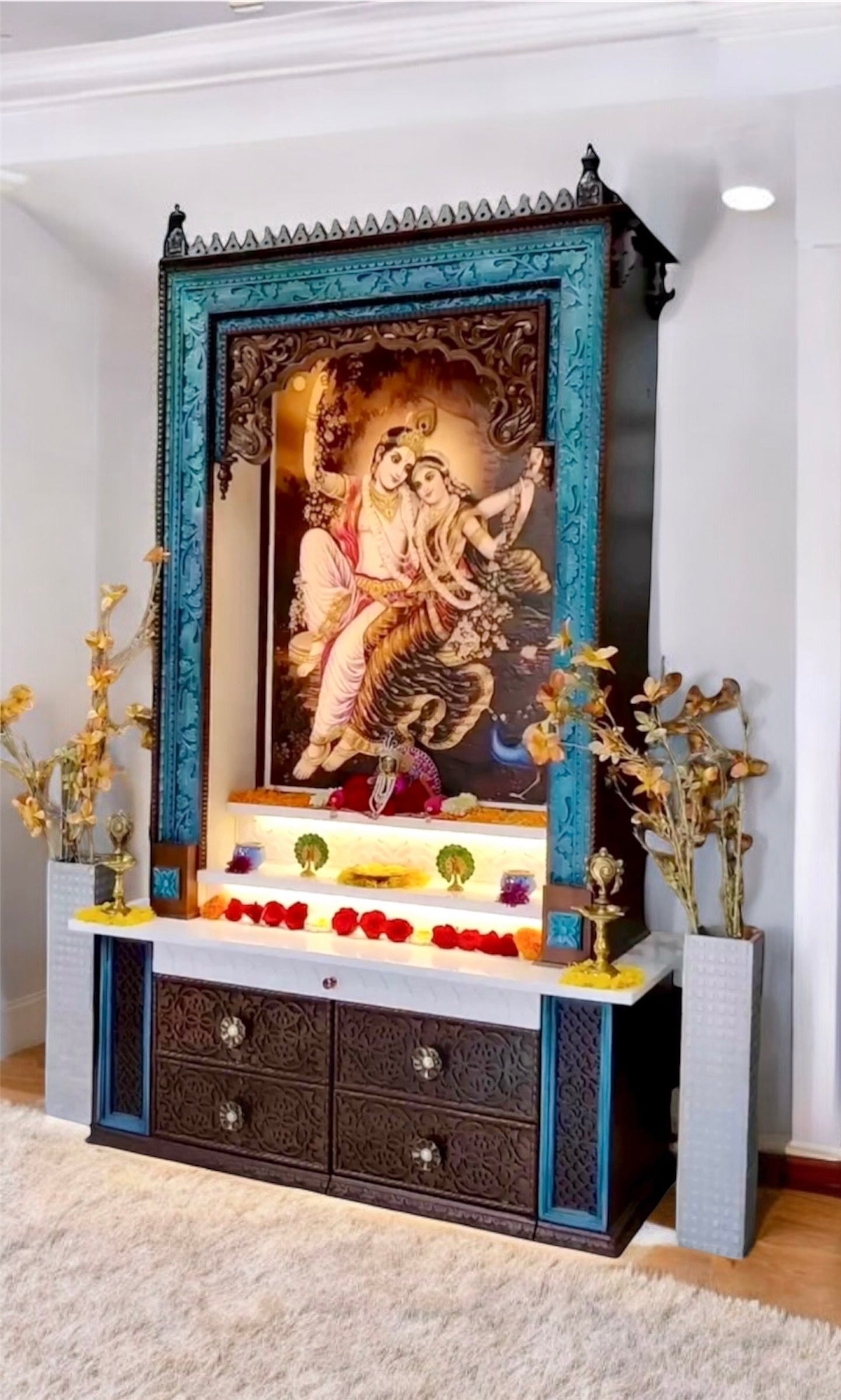 Traditional large wooden pooja mandir with a rich dark brown finish and peacock color accents, featuring a Corian picture of Radhe Krishna and finely carved drawers.