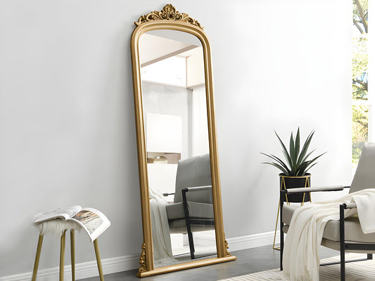 Elegant gold ornate full-length mirror leaning against a white wall in a modern living room with minimalist furniture and a potted plant.