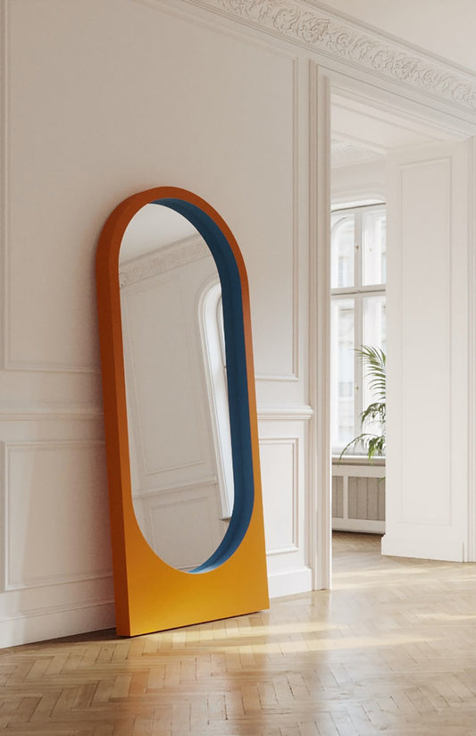 Modern arched full-length mirror with orange and blue color block frame leaning against a classic white wall in a stylish room.
