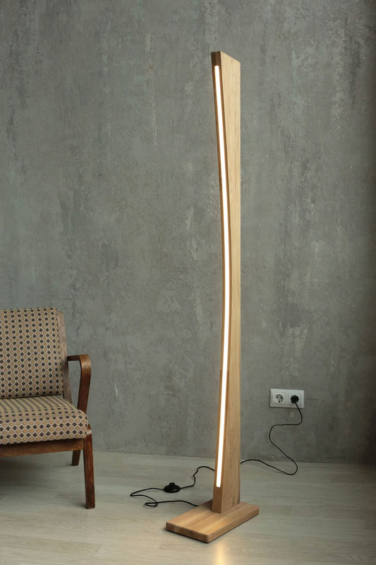 Modern curved wooden LED floor lamp with warm neutral light, standing next to a vintage upholstered chair against a textured gray wall.