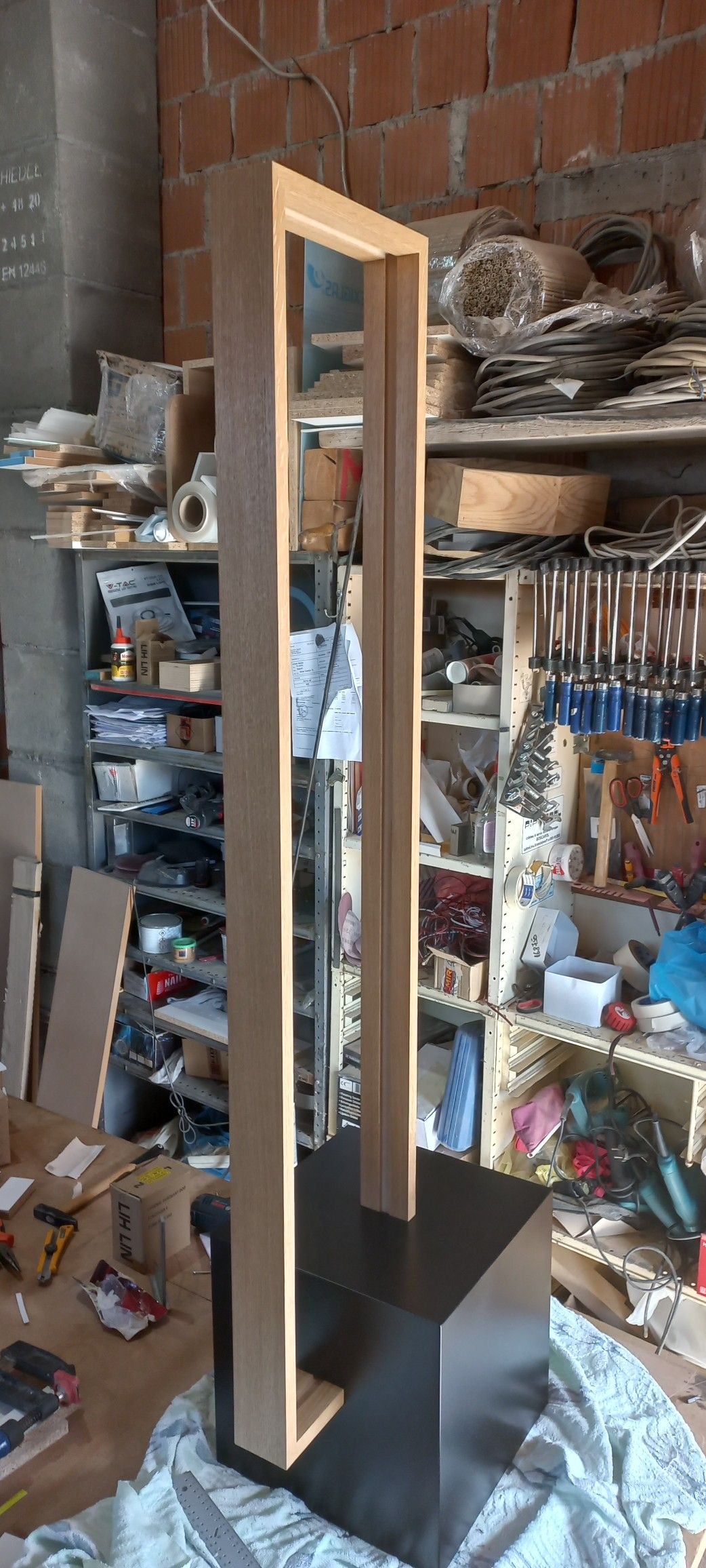 Tall wooden lamp with black cube base in a workshop setting with tools and materials in the background