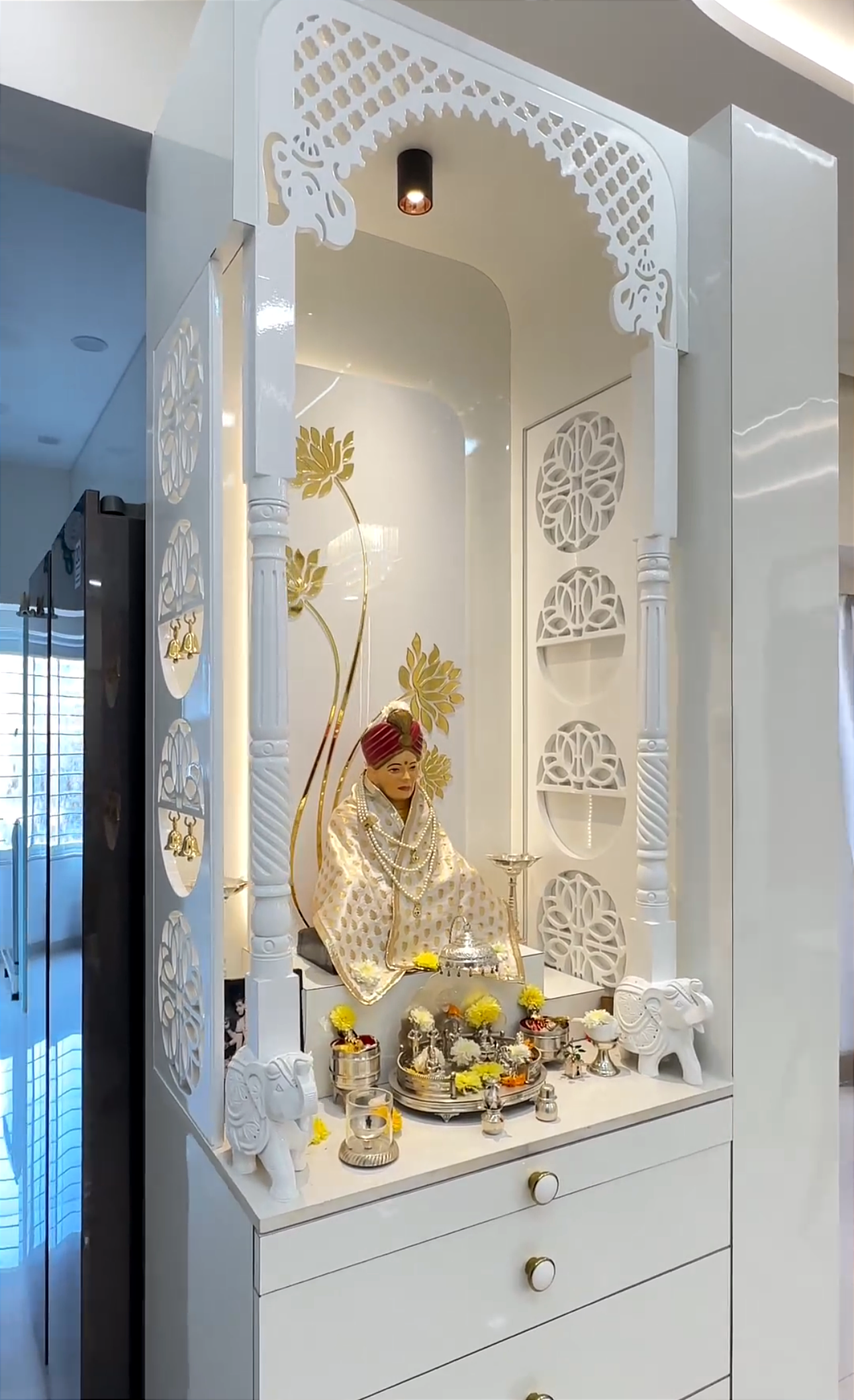 Designer white wooden Pooja mandir with hand-carved lattice work, gold floral wall decor, and silver pooja thali, creating a serene and sacred atmosphere.