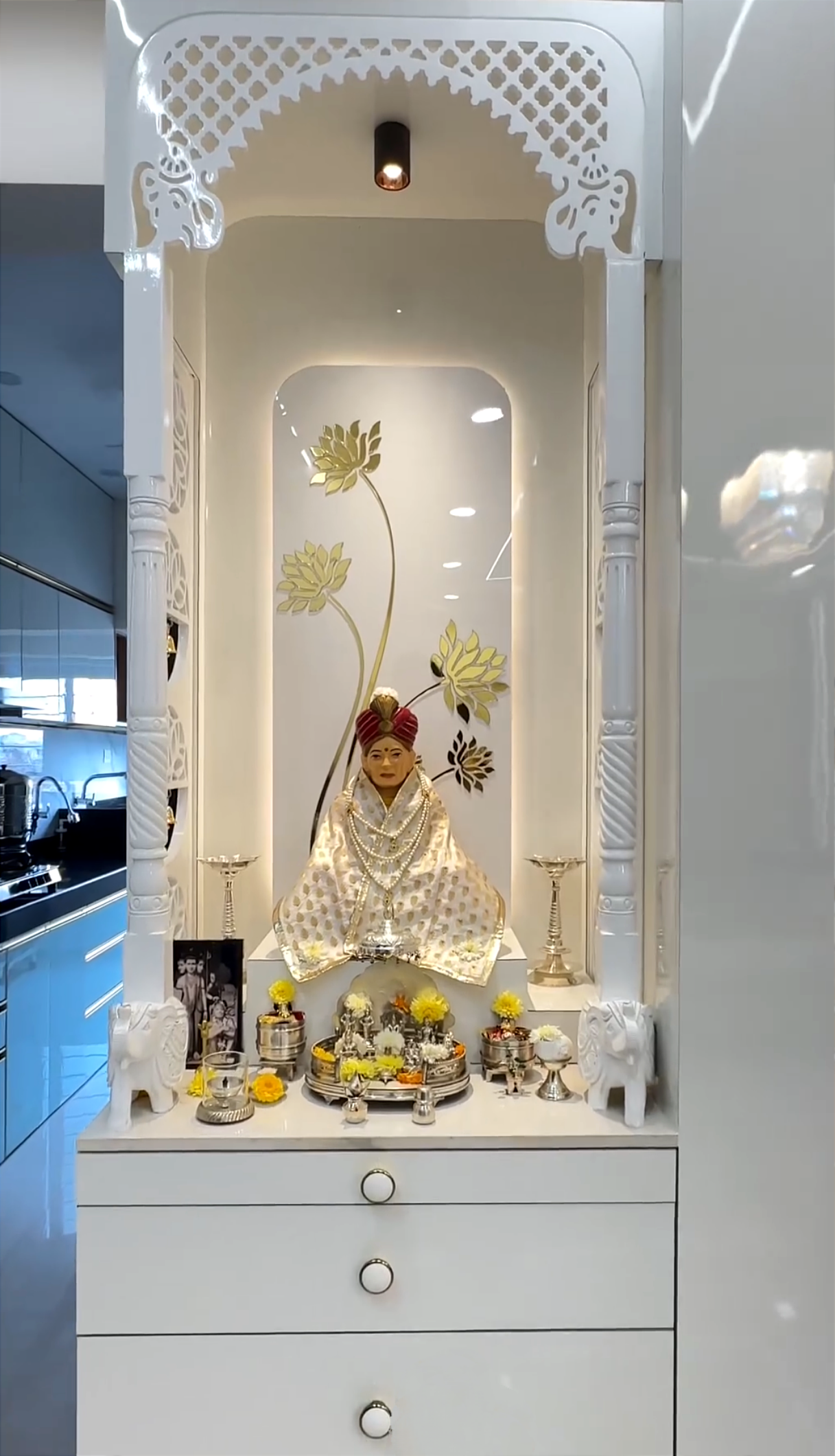 Modern white Pooja mandir with gold lotus wall design, handcrafted wooden pillars, and decorative bells, showcasing a beautifully dressed deity statue with silver pooja accessories.