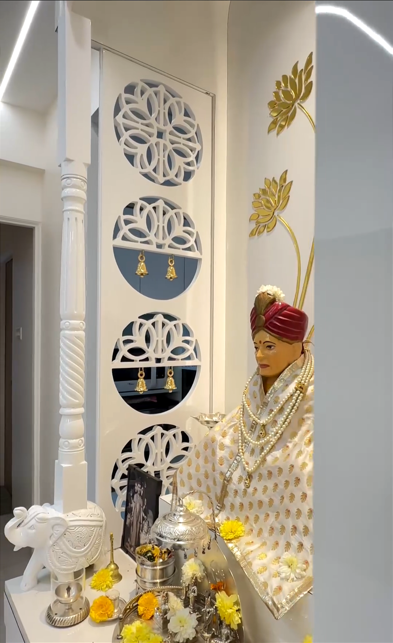 Elegant white wooden Pooja temple with intricate carvings and golden floral accents, featuring a traditional deity statue adorned with pearls and a decorative backdrop.